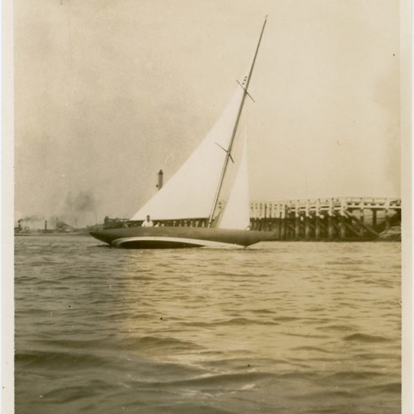 Black and white photograph of a Six Metre boat sailing past a harbour wall.
