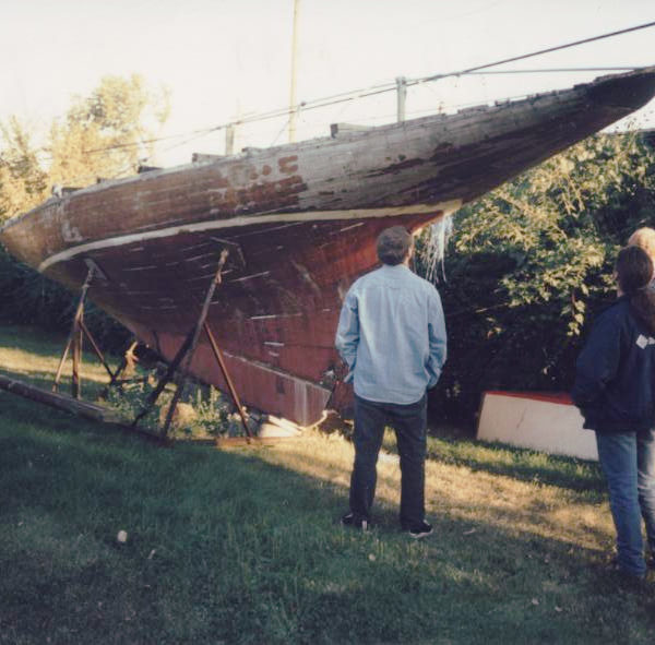 A hull of a yacht in poor condition on a support on grass. Three people are observing the hull.
