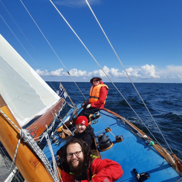 Three people sailing on a six metre boat on water with blue skies and clouds in the background
